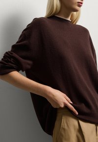 Brauner Wollpullover mit lockerer Passform, umgeschlagenen Bündchen und hohem Kragen; kombiniert mit beigen Hosen, die eine sanfte Textur und ein einfaches Design zeigen.
