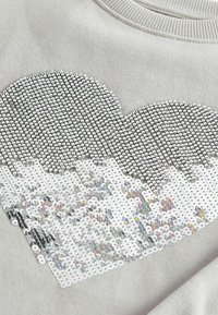 Motif de cœur en sequins argentés et blancs sur un fond de tissu gris clair, gros plan montrant la texture et les détails des coutures.