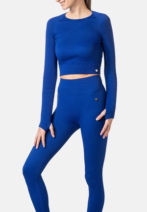 Frau trägt ein eng anliegendes, langärmeliges, königsblaues Crop-Top und passende hoch taillierte Leggings vor einem einfarbigen Hintergrund.