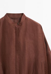 Chemise en lin marron avec col mandarin, un seul bouton supérieur, et patte de boutonnage dissimulée sur un fond blanc.