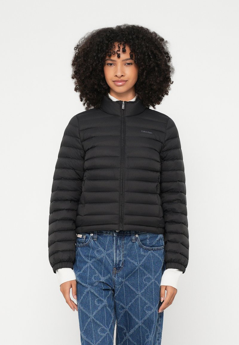 Γυναίκα με μαύρο μπουφάν με φερμουάρ και καπιτονέ υφή πάνω από λευκά μακριά μανίκια και μπλε τζιν με σχέδια, στέκεται μπροστά σε λευκό μονόχρωμο φόντο.