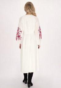 Weißes Kleid mit langen, bestickten Ärmeln, die rote Blumenmuster zeigen. Das Kleid hat eine taillierte Taille und reicht bis zur Mitte der Waden. Schwarze Stiefel.