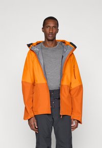 Mies, joka on pukeutunut avonaiseen kaksiväriseen oranssiin takkiin, harmaaseen pitkähihaiseen puseroon ja mustiin housuihin, seisoo yksinkertaista vaaleaa taustaa vasten.