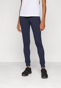 Navyblauwe, aansluitende legging gemaakt van zachte stof, met een gladde textuur en geribbelde manchetten. Draagt met een aansluitend wit shirt en zwarte schoenen.