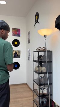 Moški z črnim duragom in zeleno majico stoji v sobi z vinilnimi ploščami na steni, talno svetilko in prozorno polico za čevlje, napolnjeno s čevlji.