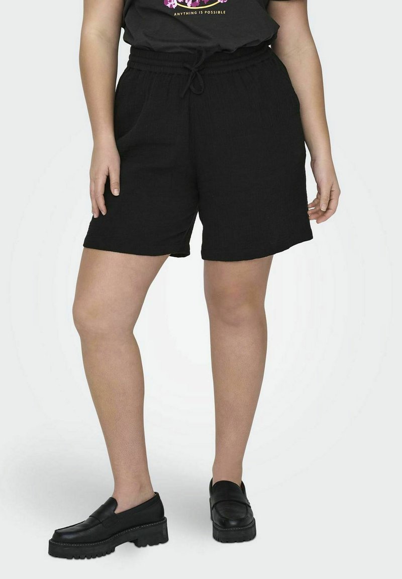 Shorts noirs en tissu texturé, dotés d'une taille élastique avec cordon de serrage et de poches latérales. Portés avec des chaussures plates noires.