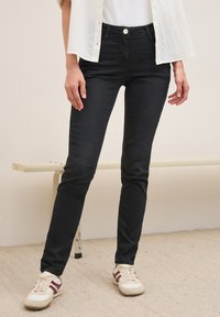 Jeans negros de corte ajustado, con cierre de botón en la parte delantera y presillas para cinturón. Combinados con zapatillas blancas y burdeos sobre un suelo texturizado.