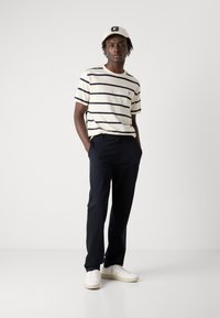 Stribet creme og marineblå T-shirt med korte ærmer, marineblå bukser, hvide sneakers og en lys farvet kasket med et logo.