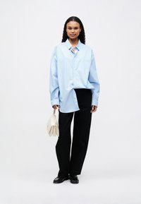 Camisa oversized azul e branca às riscas, calças largas pretas e uma mala crème, com uma textura suave e detalhes em botão.