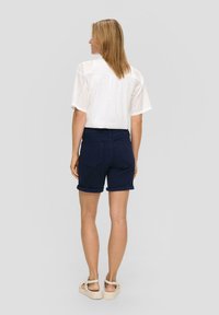 Witte blouse met oogjes en korte mouwen, marineblauwe shorts met opgerolde mouwen en beige sandalen. Aansluitend ontwerp met achterzakken op de shorts. Gladde texturen.