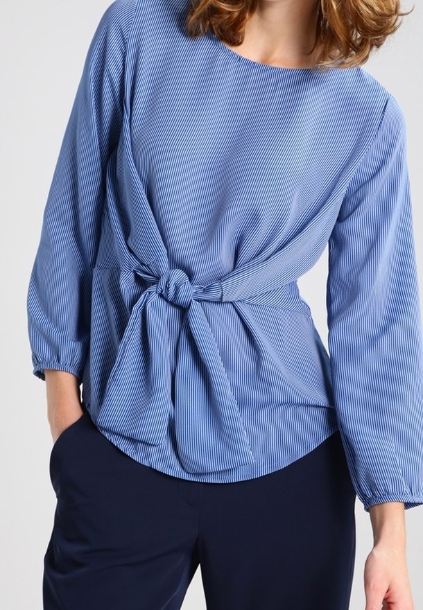 Femme portant un chemisier bleu à rayures fines à manches longues, noué à l'avant à la taille, et un pantalon bleu marine foncé.