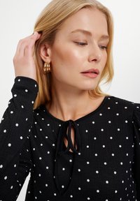 Czarny top w kropki, z długimi rękawami, detalem w postaci wiązania pod szyją i teksturowanym materiałem. Podkreślony przez modelkę noszącą złote kolczyki w kształcie kół.