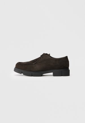 Donkerbruin suède veterschoen met een zwarte chunky rubberen zool, moc toe stiksels en een lage hak, getoond vanaf de binnenzijde tegen een eenvoudige achtergrond.