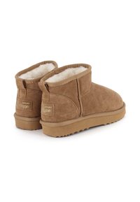 Botas de ante marrón hasta el tobillo con forro de pelo sintético, suela plana de goma y pequeño detalle del logo en la parte trasera, con punta redondeada y bordes cosidos.