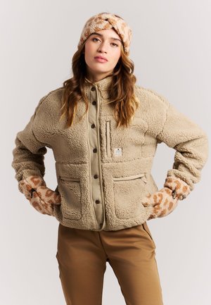 Woman wearing a beige sherpa jacket, matching leopard print fleece gloves, headband, and brown pants, standing with hands on hips.