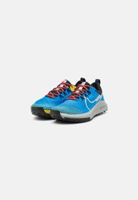 Scarpe da ginnastica blu con accenti neri, parte superiore in mesh, suola texturizzata, lacci rossi e dettagli riflettenti. Presentano un'etichetta gialla distintiva.