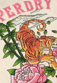 Okraski dizajn rjovečega tigra, obdani z zelenimi listi, rožnatimi cvetovi in bodečo žico ter napisom "PERDRY" zgoraj na kremni tkanini.