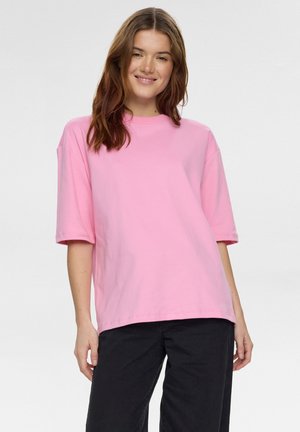 Junge Frau lächelnd, trägt ein lockeres rosa Kurzarmhemd und schwarze Hose, steht vor einem schlichten hellen Hintergrund.