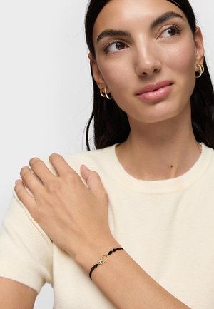 Donna con capelli scuri che indossa una camicia crema e orecchini a cerchio dorati, che mostra un braccialetto con cordino nero e un ciondolo ovale dorato al polso.