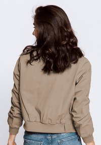 Beige bomber jacket with ribbed cuffs and hem. Smooth, lightweight fabric with a relaxed fit, shown from the back. Blue denim jeans visible.