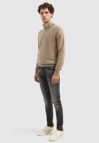 Man met steil haar in een beige sweater met kwartrits, vervaagde zwarte jeans en witte sneakers, staand tegen een lichtgrijze achtergrond.