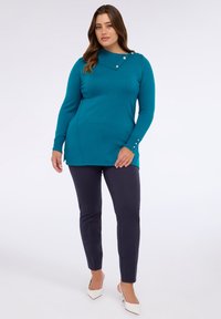 Teal långärmad topp med asymmetrisk neckline och två knappdetaljer, i kombination med marinblå byxor och spetsiga vita klackar. Slät textur, figurnära stil.