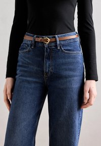 Cureau din piele maro cu cataramă circulară aurie purtat peste blugi de culoare albastru închis. Blugii au o croială dreaptă și un design clasic cu cinci buzunare.