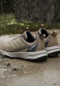 Beige udendørssko med mesh- og syntetiske materialer, tekstureret design, gummisål og en trækkant; har 'TERREX' branding i grå.