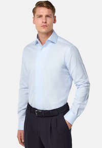 Chemise à manches longues rayée bleu clair et blanche avec un col classique, devant à boutons, et une ceinture tissée noire texturée sur un pantalon sombre.