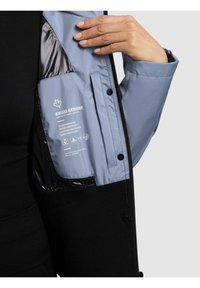 Mano che apre una giacca nera e azzurro chiaro mostrando l'etichetta interna con caratteristiche di impermeabilità, idrorepellenza, leggerezza e protezione dal vento.