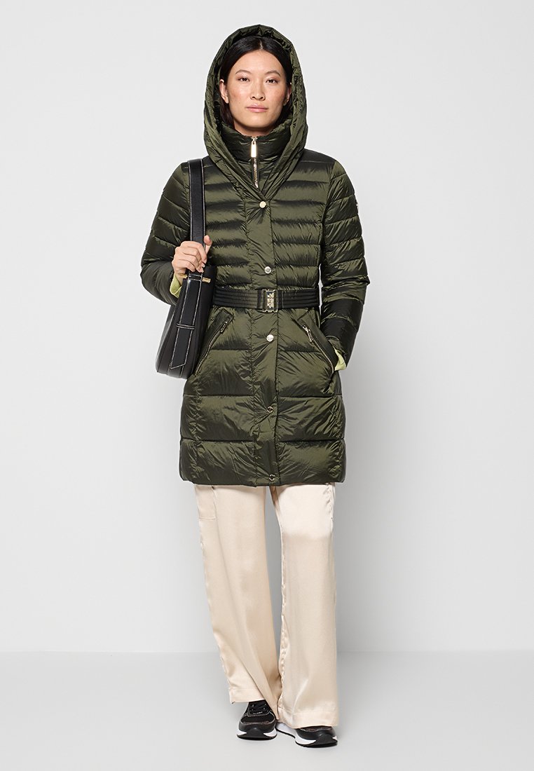 Frau in einem olivgrünen, mit Gürtel versehenen Puffermantel mit Kapuze, cremefarbenen weiten Hosen, schwarzen Sneakers und einer schwarzen Umhängetasche.