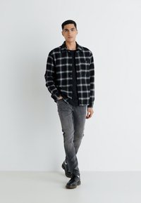 Chemise à carreaux noir et blanc, portée sur un t-shirt noir ; jean gris et chaussures noires. Le mannequin se tient sur un fond clair.