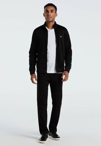 Zwarte zip-up jas met groene zijaccenten, gedragen over een witte t-shirt. Slim-fit zwarte broek en zwarte sneakers maken de outfit compleet.