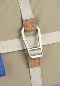 Dettaglio di una borsa beige con clip in metallo argento, accento in sughero e tracolla in tessuto bianco. Include un'etichetta in tessuto blu sul lato.