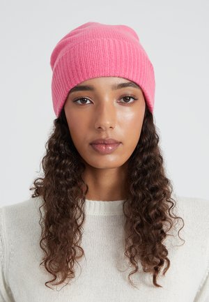 Giovani donna con lunghi capelli ricci, che indossa un berretto di maglia rosa e un maglione di maglia bianco, che guarda in avanti davanti a uno sfondo neutro.