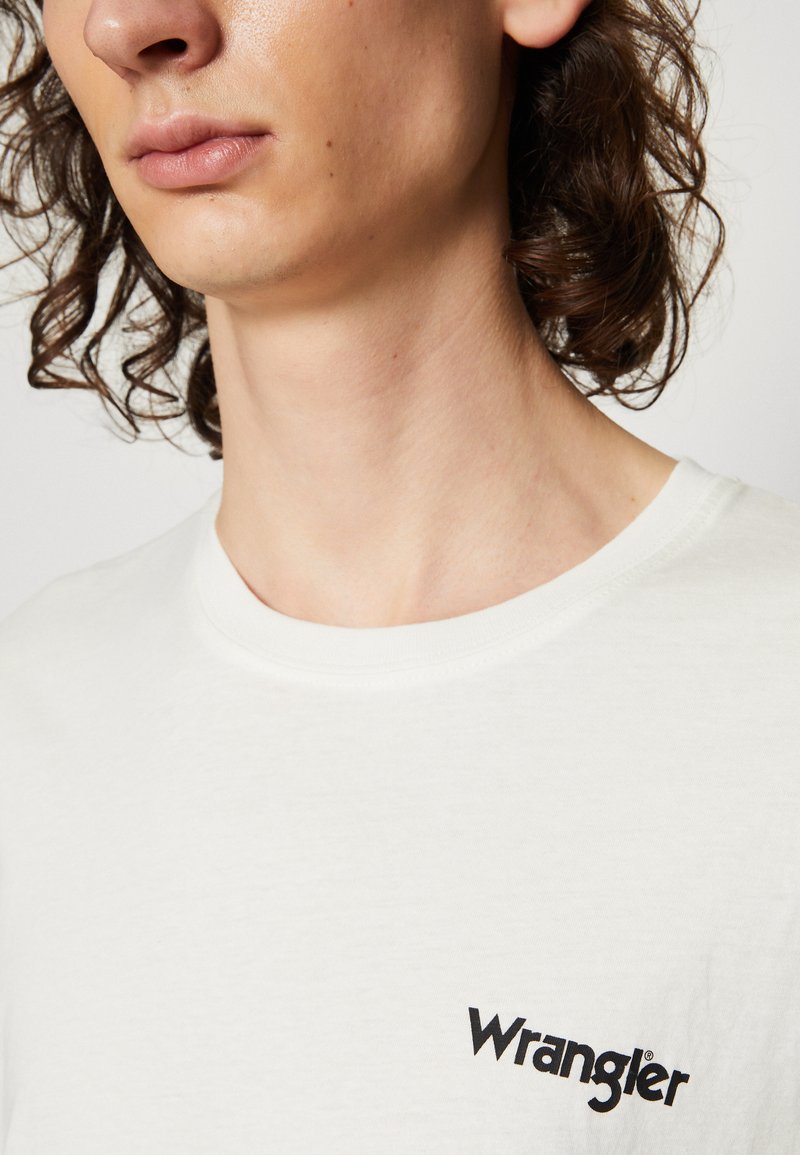 T-shirt di cotone bianco con colletto a girocollo e logo "Wrangler" nero sul lato sinistro del petto. Texture liscia con un design minimale.