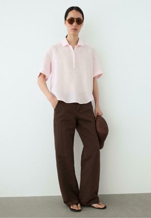 Femme portant un t-shirt rose clair à manches courtes, pantalon large marron, tongs, lunettes de soleil, tenant un chapeau tressé marron contre un mur blanc uni.