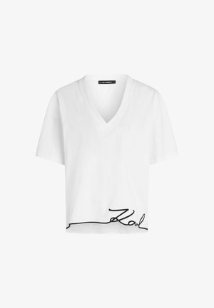 Witte cropped t-shirt met een v-halslijn en korte mouwen. Heeft een zwarte cursieve handtekening aan de onderrand. Zachte stoftextuur.