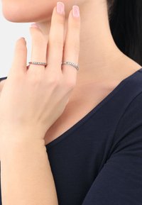 Två ringar bärs på fingrarna; en är en enkel silverring, medan den andra har små klara stenar, båda med en elegant design.