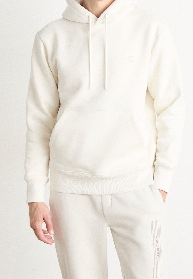 Personne portant un sweat à capuche blanc avec une poche avant et un pantalon de survêtement blanc assorti avec le logo Calvin Klein sur la cuisse gauche.