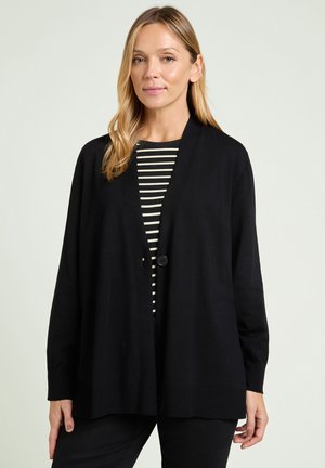 Woman wearing a loose black cardigan over a black and white striped shirt, standing against a plain light background.