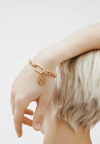 Pulsera de cadena de oro con un gran broche, que presenta un charm decorativo con letras abstractas. Textura suave y brillante. Tono de piel visible debajo.