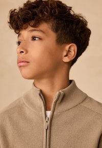 Ragazzo con i capelli ricci guarda di lato, indossando un maglione beige con zip su uno sfondo beige semplice.