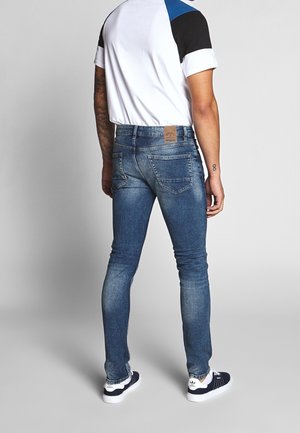 Mand iført blå slim-fit jeans, hvid og sort kortærmet skjorte og sorte og hvide sneakers, stående med ryggen mod seeren.