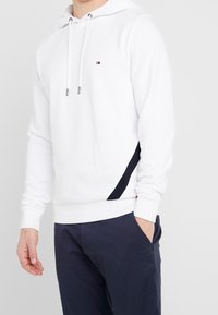 Man in een witte hoodie met een klein logo en marineblauwe broek, met één hand in zijn zak tegen een effen lichte achtergrond.