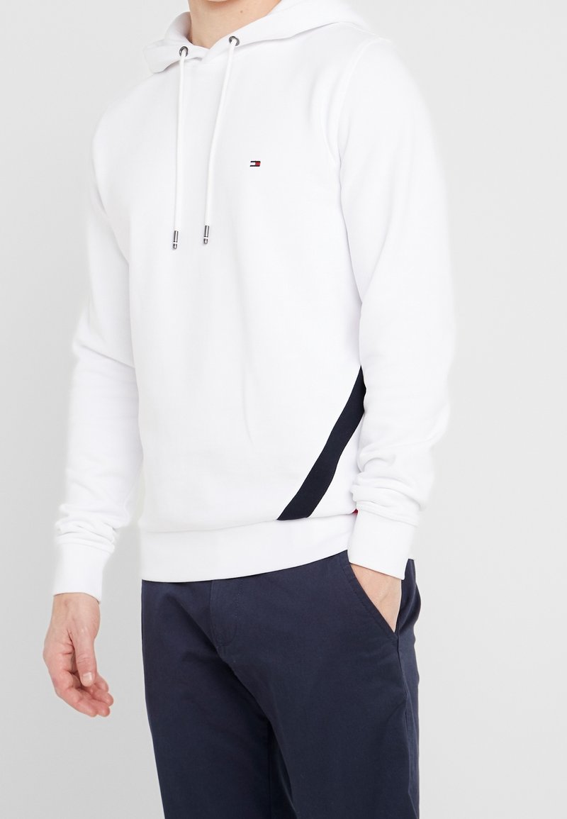 Man in een witte hoodie met een klein logo en marineblauwe broek, met één hand in zijn zak tegen een effen lichte achtergrond.