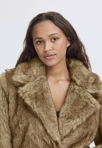 Giacca in pelliccia sintetica di colore marrone chiaro, con un ampio colletto, morbida texture e vestibilità ampia. Ideale per calore e comfort in condizioni di freddo.