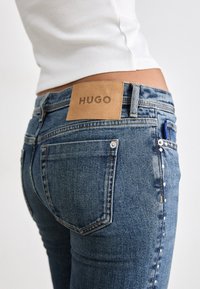 Siniste pesemisega teksapüksid, millel on pruuni nahast silt, kuhu on pressitud "HUGO", ja metallist nurgarõngad külgedel. Kitsas lõige.