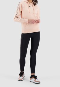 Felpa rosa chiaro con logo sul petto, cappuccio con lacci e strisce nere di accento. Abbinata a leggings neri e sneakers sportive multicolori.