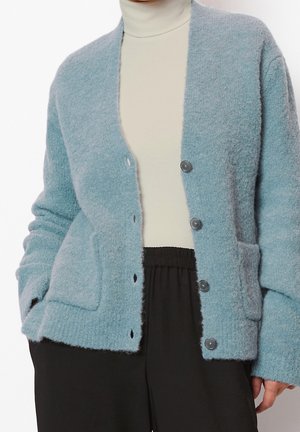 Hellblauer, flauschiger, geknöpfter Cardigan, getragen über einem cremefarbenen Rollkragenpullover und kombiniert mit schwarzen Hosen mit elastischem Bund.